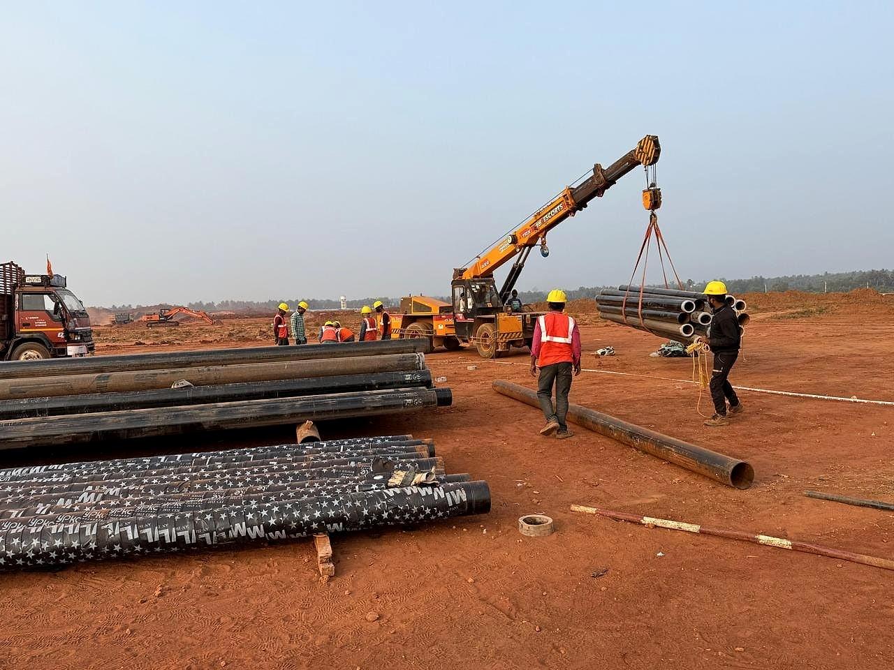 Crane lowering a steel pipe as a banksman in PPE guides the load, Bhogapuram International Airport